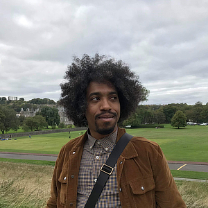 You are shown a photograph of a man. His skin: brown, like
                            the bark of a tree; his hair: black and coiled like springs,
                            except for a few white wiry strands. He is wearing a
                            chocolate brown corduroy jacket over brown and white
                            checkered shirt, and over his left shoulder, a black nylon
                            strap across his torso. It is obviously for some sort of bag.
                            He is smiling slightly and looking askance to something on
                            his left.